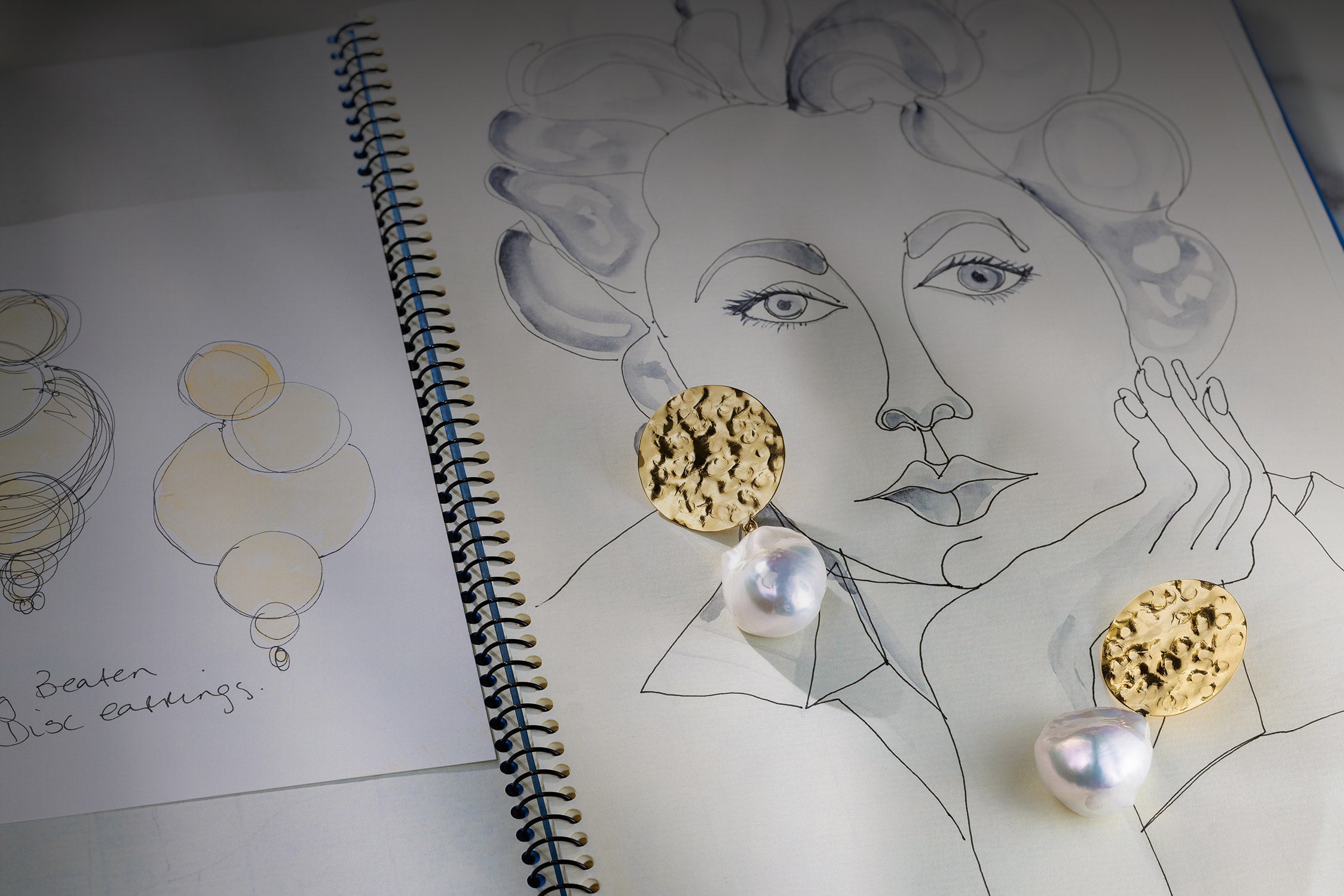 Baroque Pearl Sun Disc Earrings on a line drawing of a face with a notebook in the background by Isabella Roux