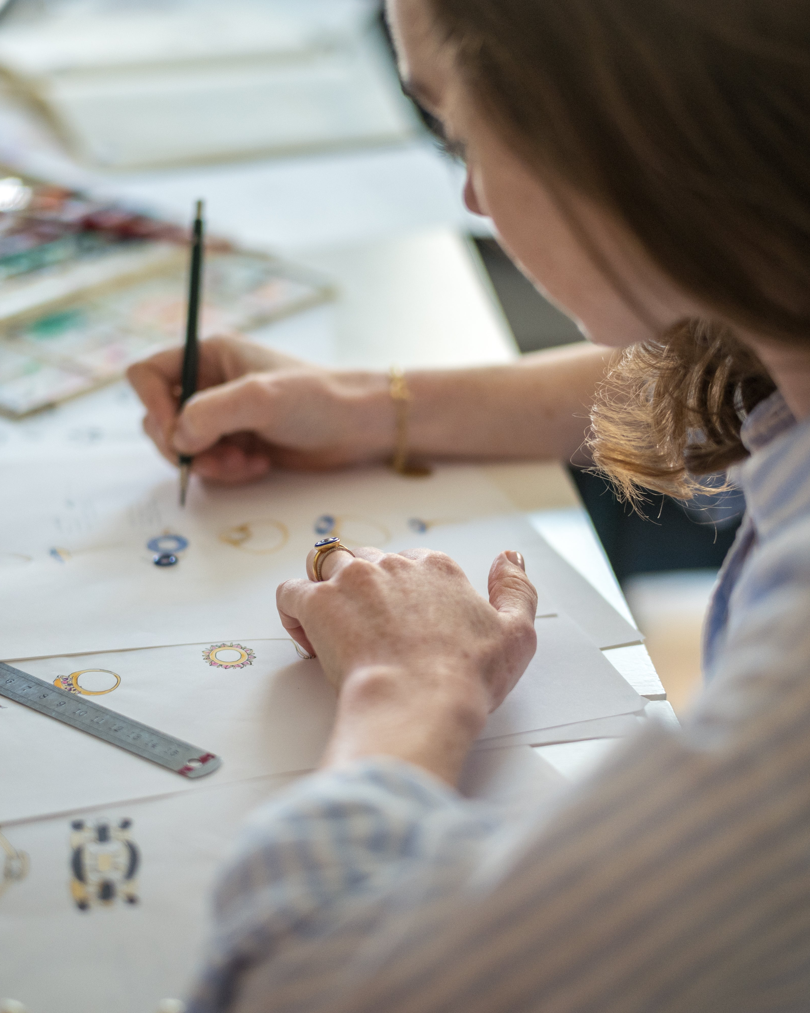 Sadhbh Isabella Roux-Fouillet, Creative Director of Isabella Roux, drawing jewellery designs on a piece of paper with a pencil, surrounded by art supplies.