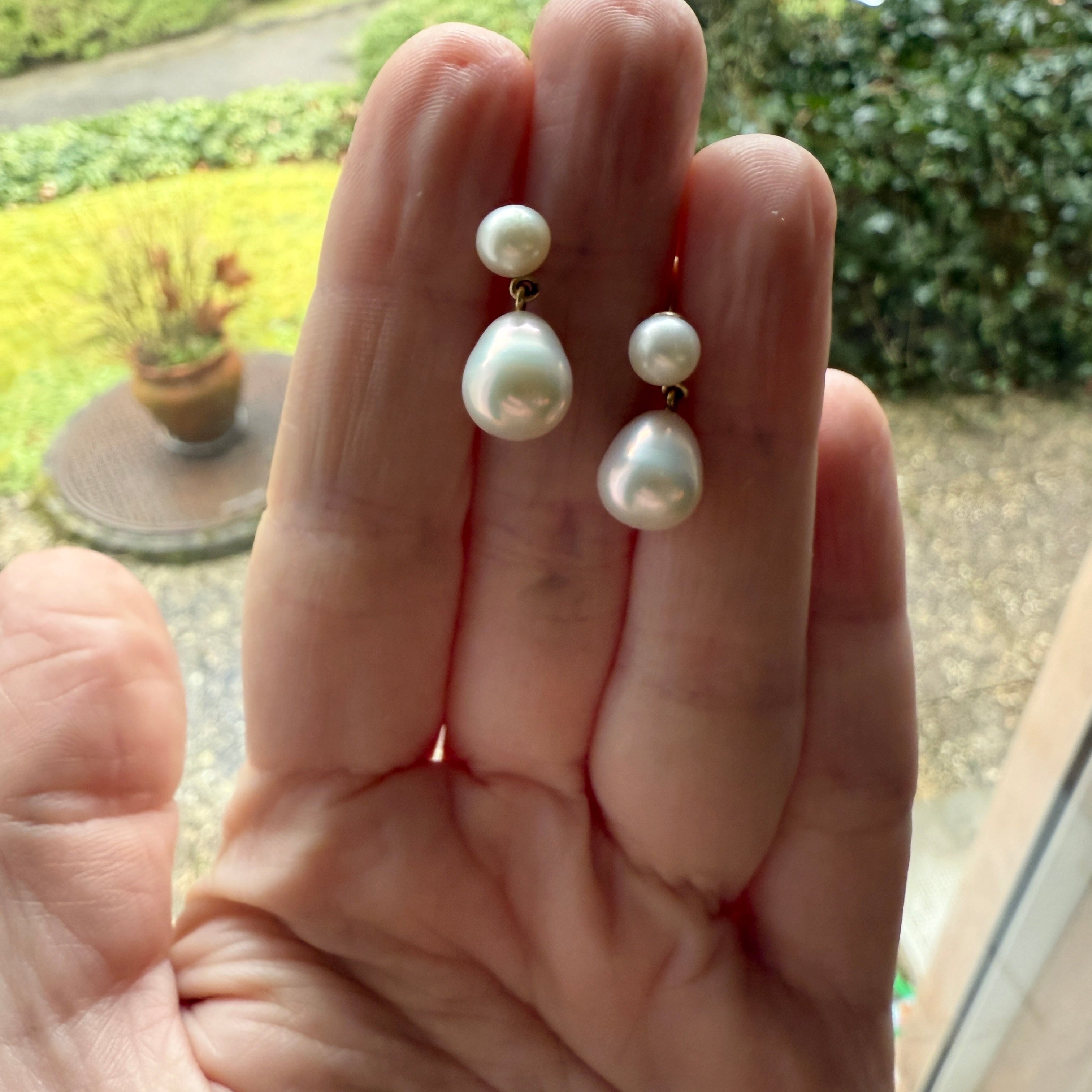 Hand showing 18K yellow gold Pearl Drop Earrings from the Pebbles Collection featuring white freshwater round and teardrop pearls by Isabella Roux.