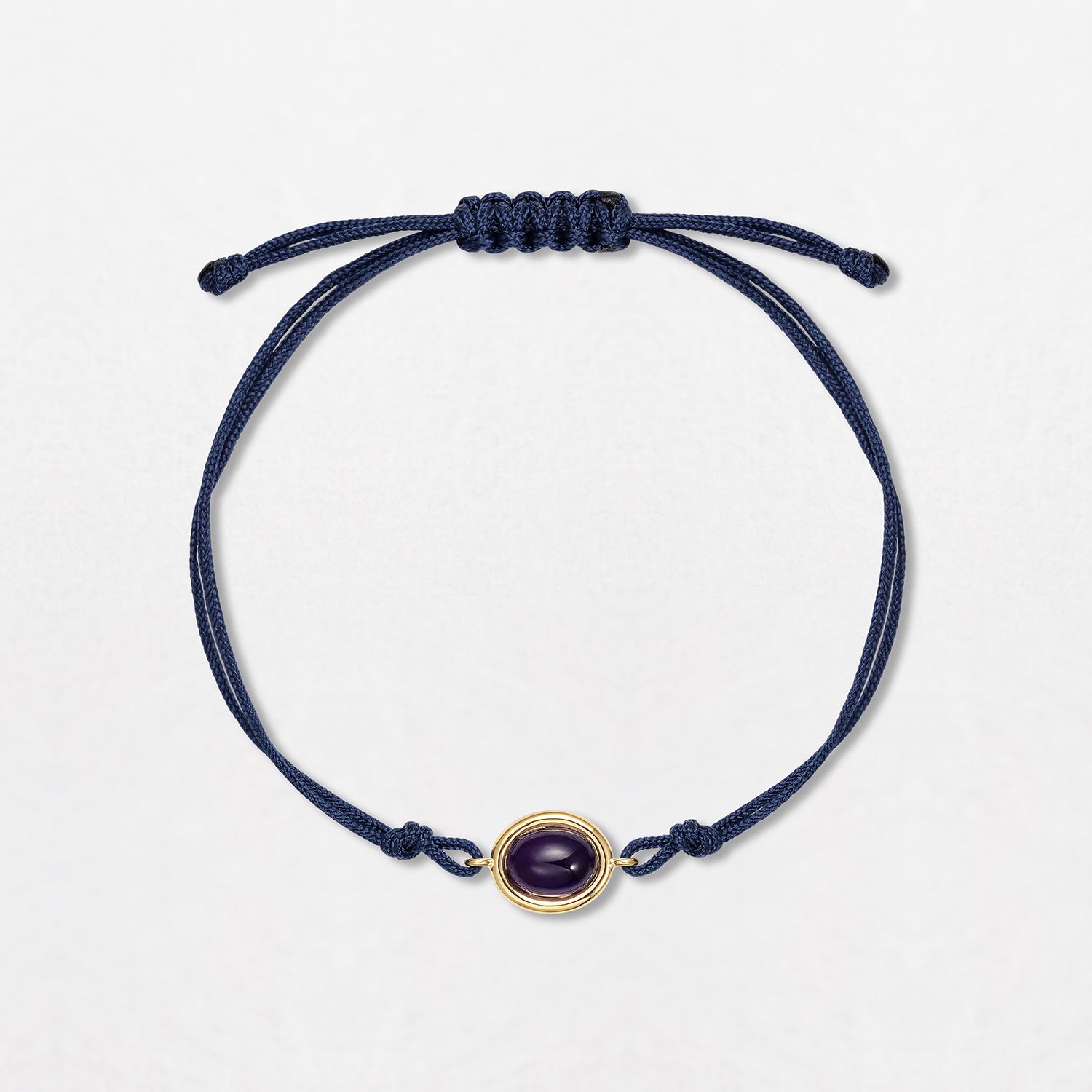 An Amethyst Pebble Bracelet with a 2ct oval amethyst cabochon, set in 18K yellow gold on a double navy blue cord. One size with an adjustable slider fastening.