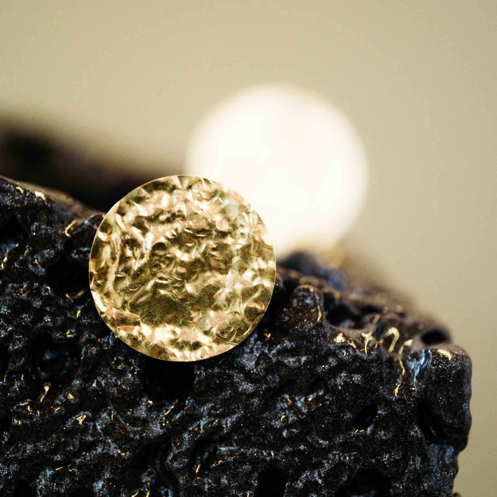 18k gold beaten earring on a textured black surface with a blurred light source in the background by Isabella Roux