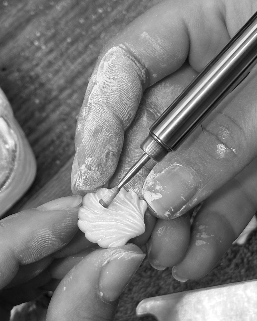 Close-up of hands using a tool on a gemstone to carve a shell shape, by Isabella Roux