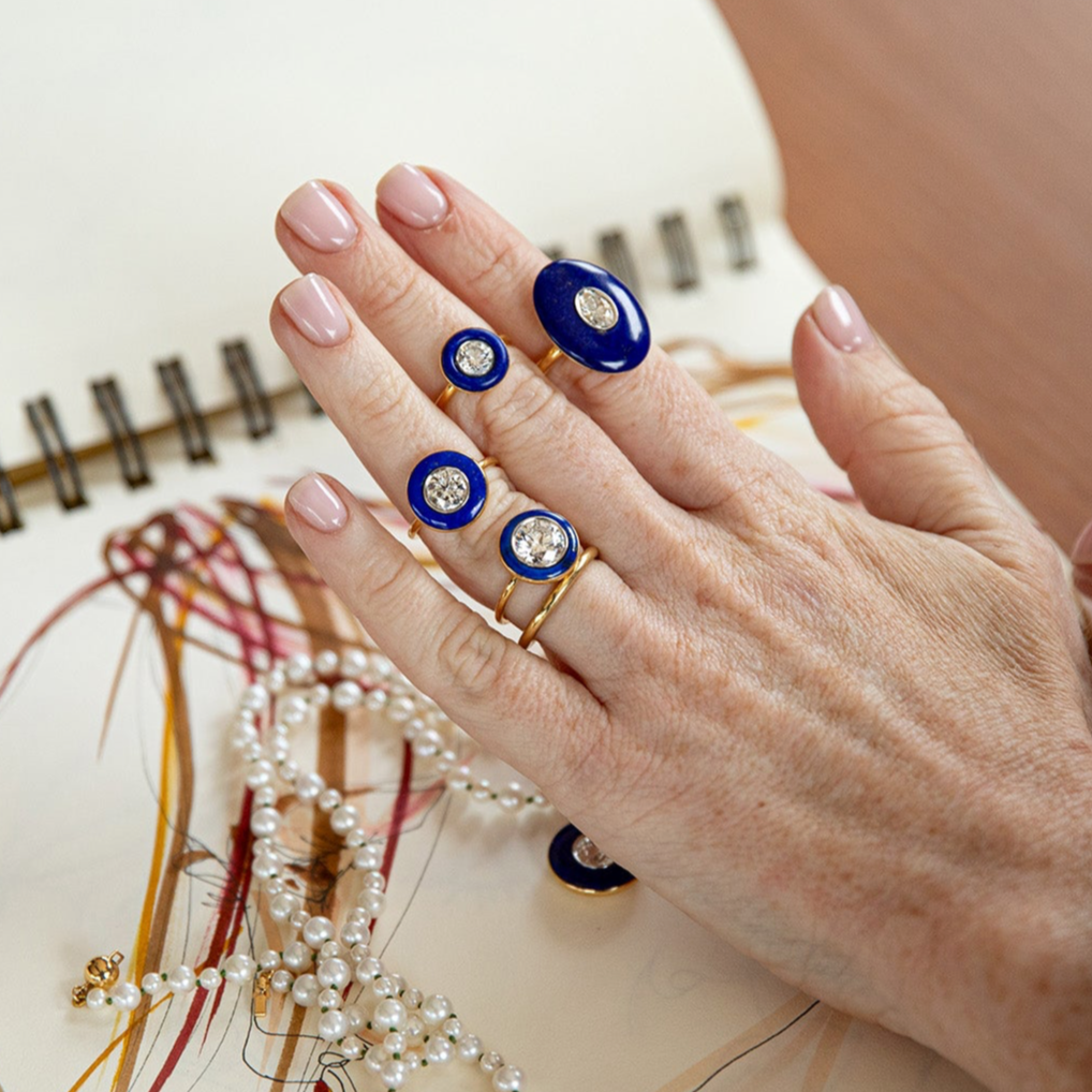 Hand wearing multiple rings with blue and gold accents on a light background. Bespoke 18K yellow & white gold Diamond & Lapis Classic Ring featuring a 2.2ct round brilliant lab grown diamond and carved deep blue lapis lazuli in a halo around by Isabella Roux.